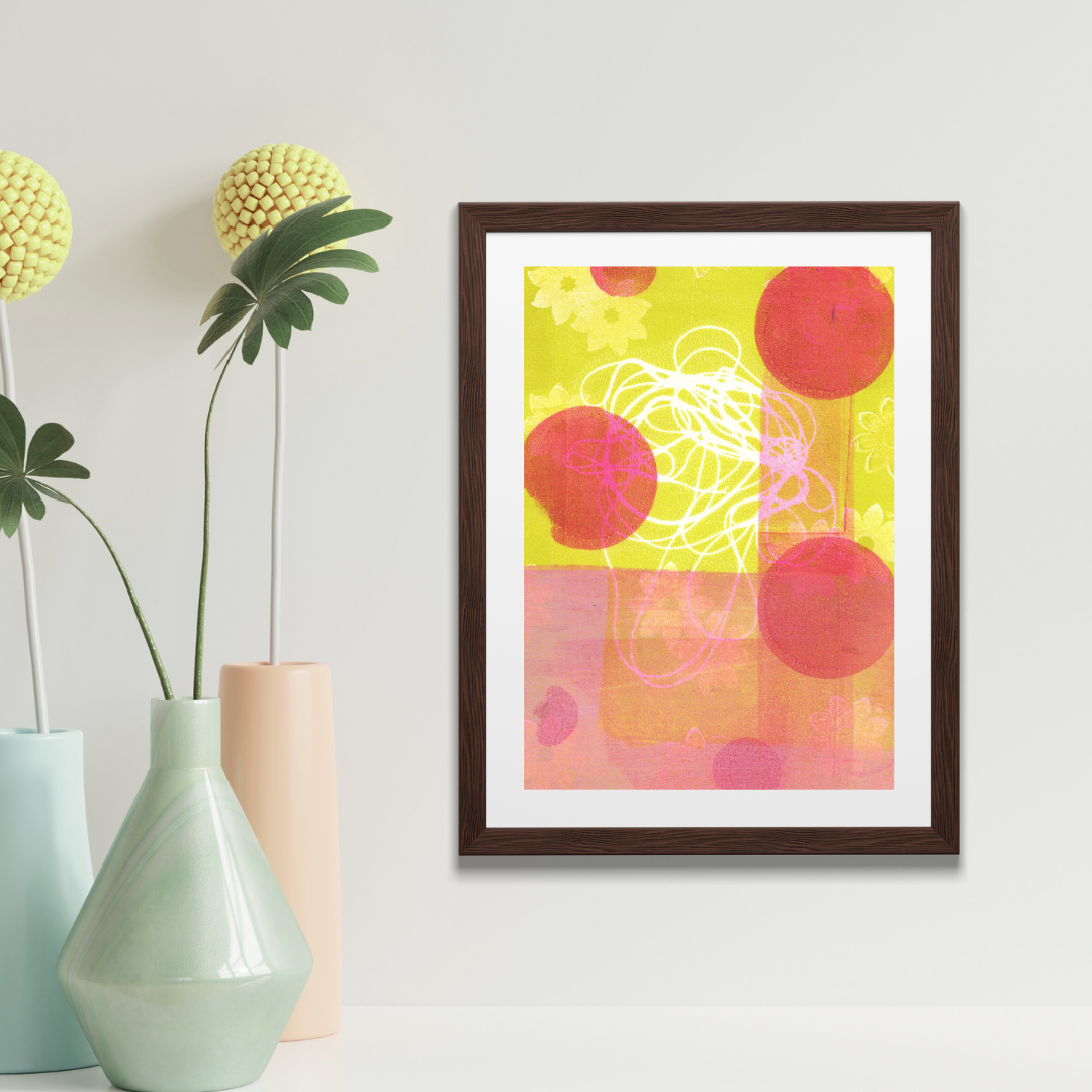 Framed abstract art print with pink circles on a white wall above a wooden surface with a plant.